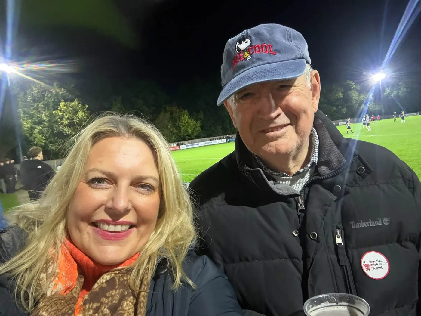 Mims Davies MP takes a selfie with a man in a cap and a Timberland jacket wearing a Cardiac Risk in the Young badge, the floodlit pitch and players behind them