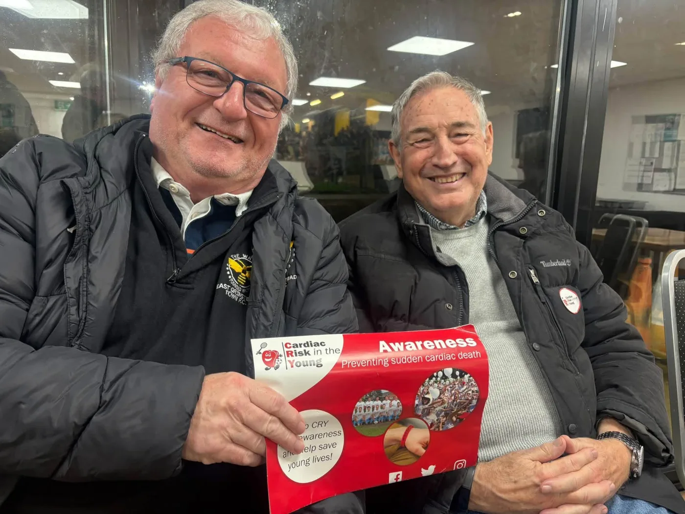 Two smiling men in padded jackets sit in the clubhouse, one holding a red Cardiac Risk in the Young awareness leaflet reading Preventing sudden cardiac death