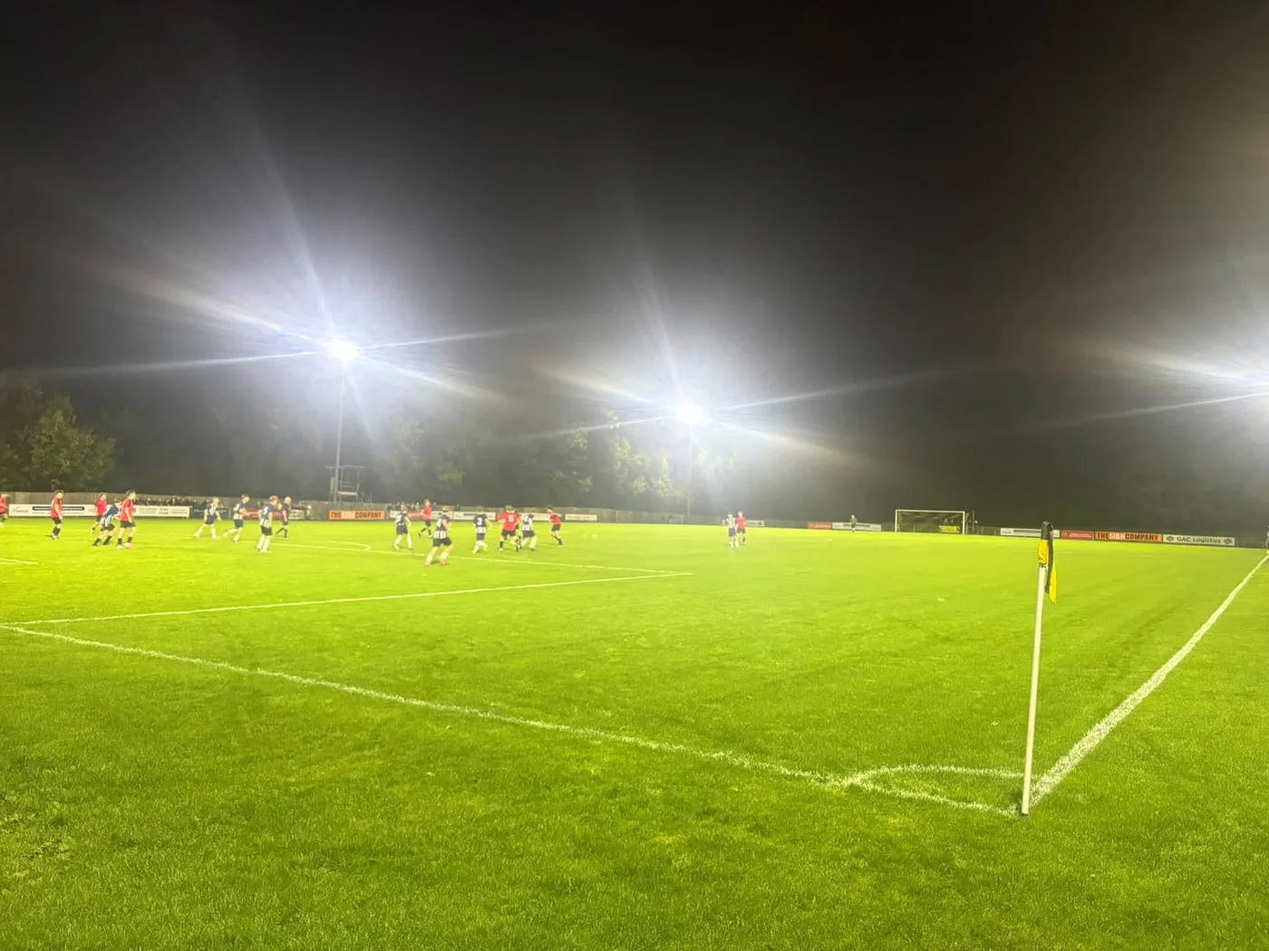 Players in red and in dark kits on a floodlit pitch at night, a corner flag in the foreground