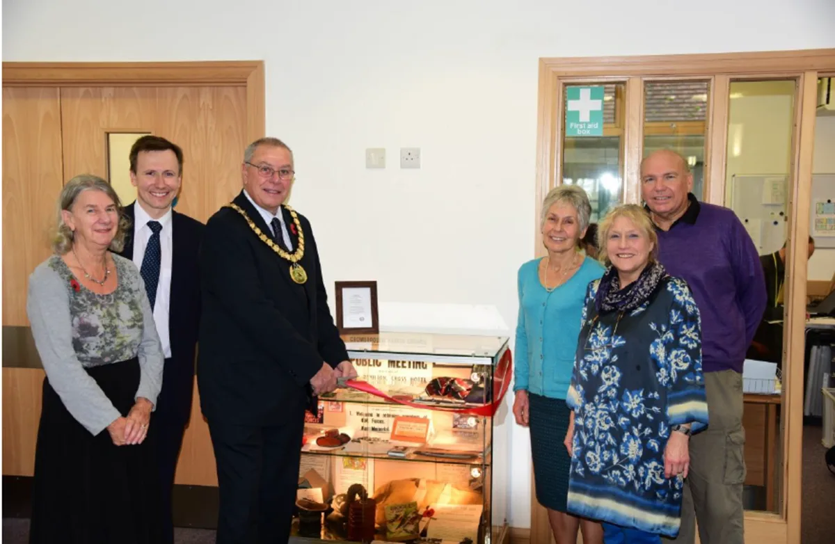 Six people stand either side of a glass display cabinet with a red ribbon across it, the Mayor in his chain of office holding scissors, in a room at Crowborough Community Centre
