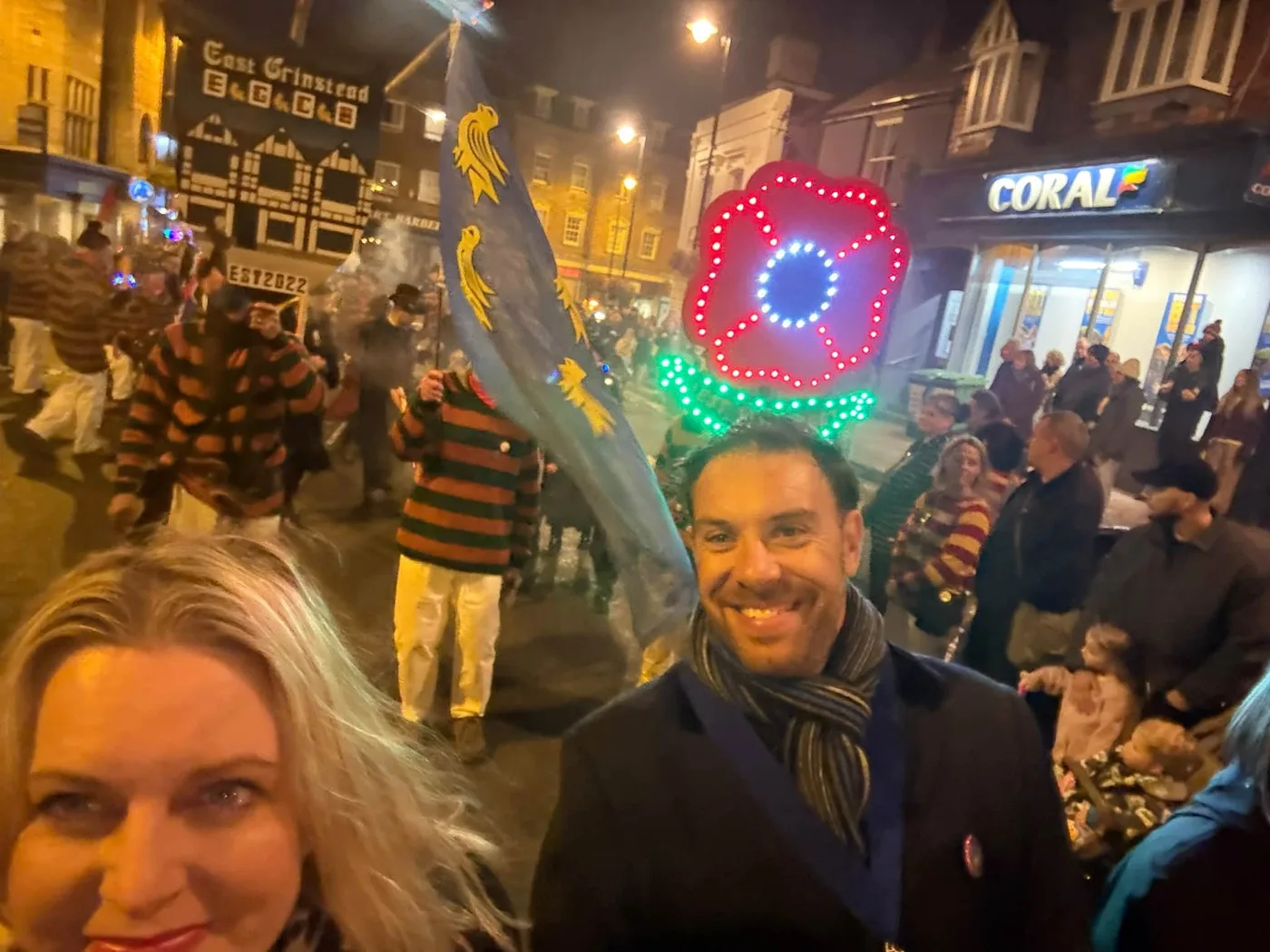 Mims Davies MP takes a selfie with a man in a dark coat and scarf as bonfire society members in striped jumpers march past with a flag and an illuminated poppy, beneath an East Grinstead ECBC banner