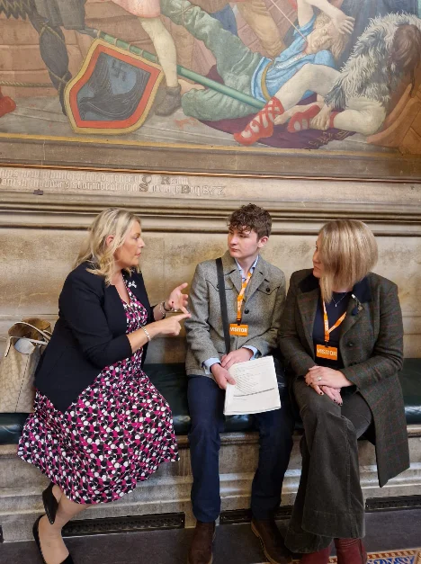 Mims Davies MP talking with her two visitors from Uckfield, who wear orange visitor lanyards, on a bench beneath a mural in the Palace of Westminster