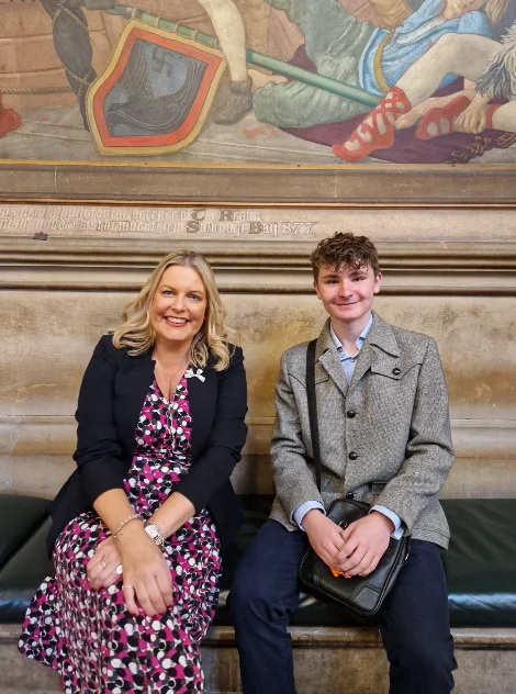 Mims Davies MP sitting on a bench with one of her Uckfield visitors in the Palace of Westminster, a mural on the wall above